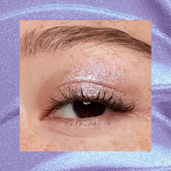 Close-up of an eye area with NYX Professional Makeup Lid Lingerie Cream Eye Pigment in 10 Lacy Lilac, shown on a duochrome blue textured background.