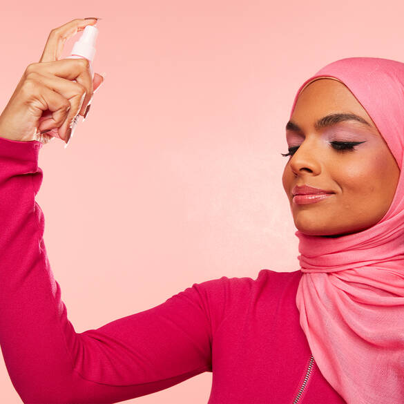 A woman in a pink hijab applies a facial spray, smiling against a soft pink background.