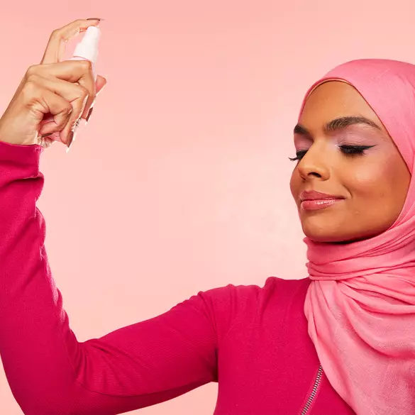 A woman in a pink hijab applies a facial spray, smiling against a soft pink background.