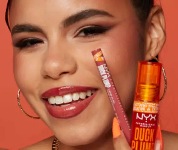 A woman smiles brightly, holding NYX lip products: a lip pencil and lip plumper, against a coral background.