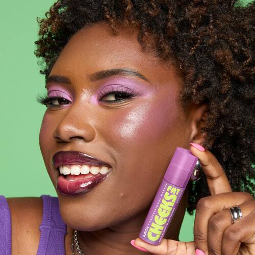 Ciemnoskora kobieta na zielonym tle prezentuje opakowanie produktu Fat Cheeks juicy blush NYX PM.