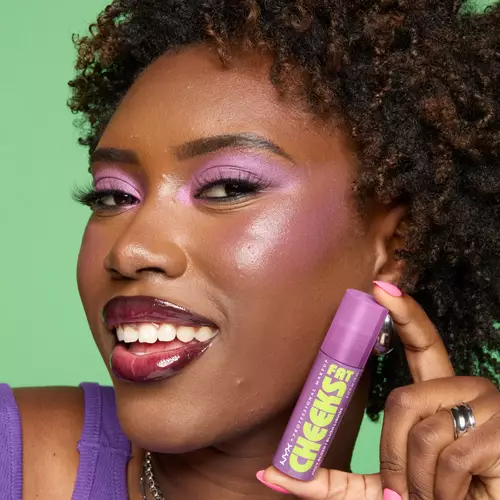 Ciemnoskora kobieta na zielonym tle prezentuje opakowanie produktu Fat Cheeks juicy blush NYX PM.