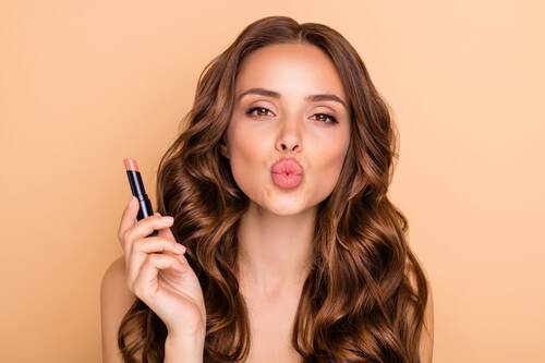 Młoda śliczna dziewczyna z rożowym błyszczykiem w dłoni przesyła całusa na pomarańczowym tle