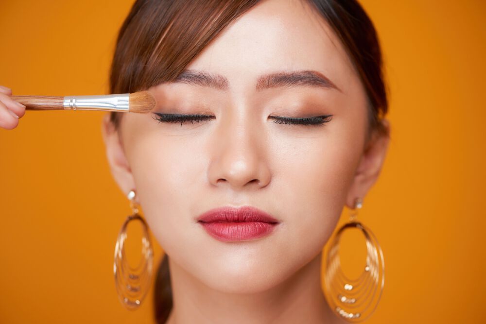 Zbliżenie na twarz kobiety o urodzie azjatyckiej, w delikatnym makijażu, ktora trzyma pędzelek do makijażu przy oku