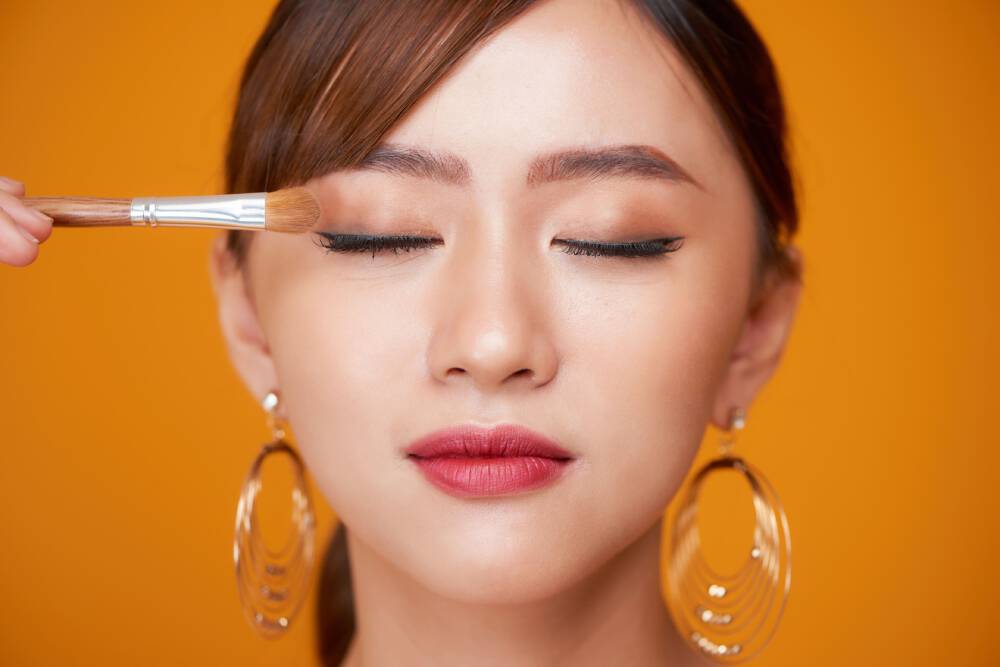 Zbliżenie na twarz kobiety o urodzie azjatyckiej, w delikatnym makijażu, ktora trzyma pędzelek do makijażu przy oku