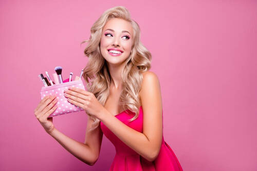 Blondynka w rożowym makeupie na rożowym tle, trzymająca w rękach kosmetyczkę z akcesoriami do makijażu