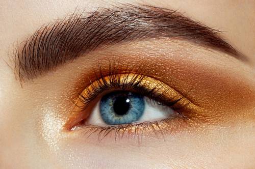 Duże zbliżenie na niebieskie oko z ze złoto-brązowymi cieniami na powiece i ułożoną brwią