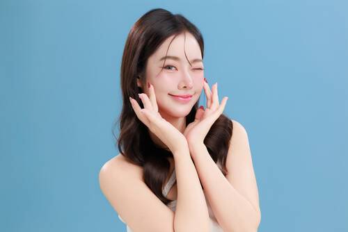 Młoda kobieta z długimi ciemnymi włosami, pozująca na niebieskim tle z dłońmi ułożonymi przy twarzy