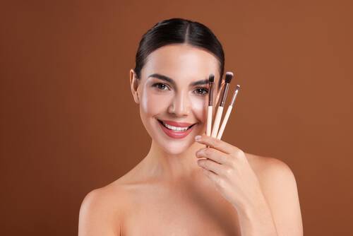 Uśmiechnięta młoda kobieta z upiętymi ciemnymi włosami, trzyma w dłoni trzy pędzle do makijażu