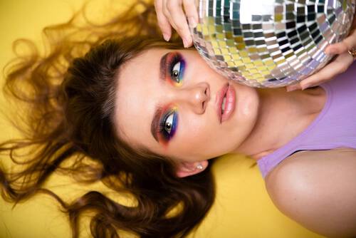 Dziewczyna na żołtym tle w mocnym kolorowym makijażu, trzymająca dyskotekową kulę