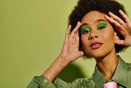 Młoda ciemnoskora kobieta w zielonej koszuli, na zielonym tle i z niebieskimi cieniami na powiekach, z dłońmi ułożonymi przy twarzy