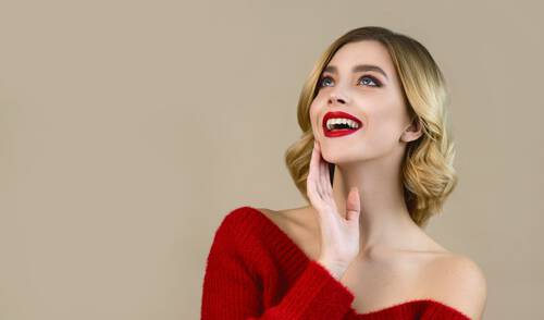 Roześmiana kobieta w pięknym makijażu i mocno czerwonych ustach dotyka dłonią twarzy i wzrok kieruje ku gorze