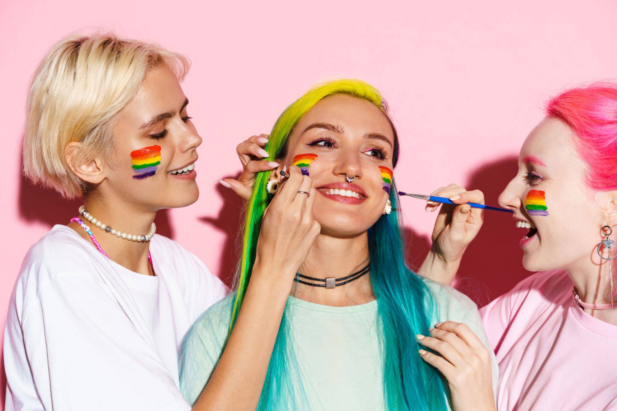 Trzy uśmiechnięte kobiety malują sobie na policzku tęczową flagę