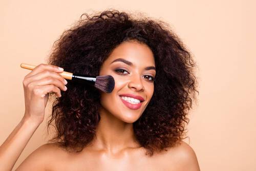 Młoda uśmiechnięta kobieta o śniadej skorze aplikuje podkład z użyciem pędzla do makijażu