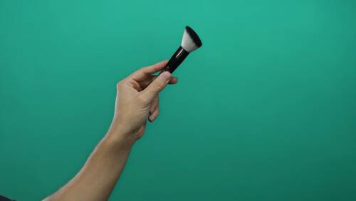 Ręka trzymająca pędzel do makijażu z czarnym trzonkiem na zielonym tle