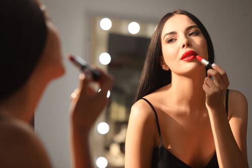 Młoda kobieta o długich włosach maluje usta czerwoną pomadką przed lustrem